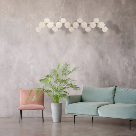 A modern living room has a gray textured wall, mint green sofa, pink velvet chair, and potted palm plant. The Dallas 25 Light Linear Suspension in gold with white glass spheres elegantly hangs from the ceiling, adding contemporary charm.
