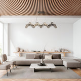 A modern living room featuring a geometrically designed chandelier, Dallas 25 Light Linear Suspension in black with smoked glass. It includes white walls, a wooden slatted ceiling, a large grey sectional sofa, a low marble coffee table, minimalist decor, and a big window enhancing the lighting.