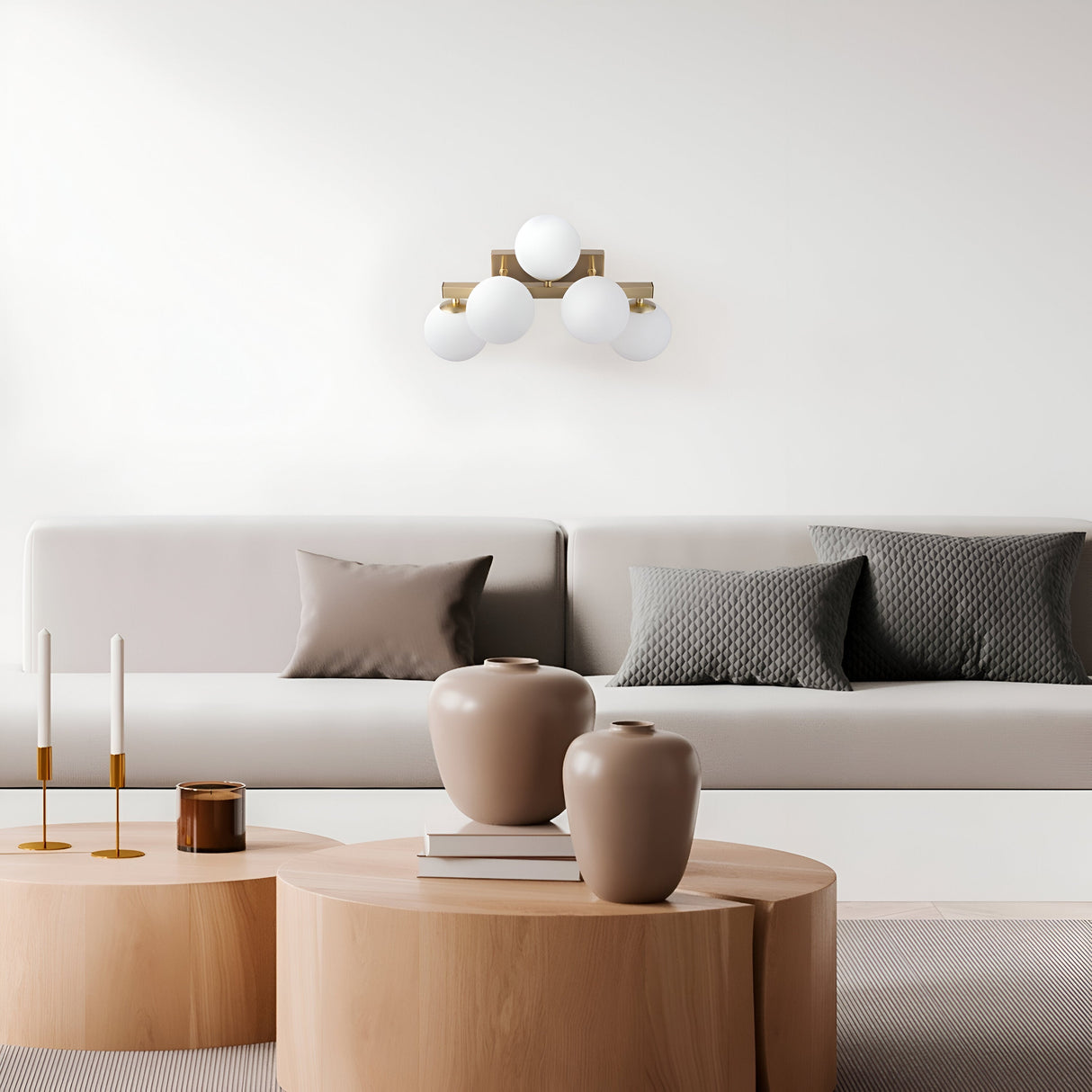 A minimalist living room showcases a long beige sofa with gray pillows. Two round wooden coffee tables hold beige vases, books, and candle holders. Above the sofa, a Dallas 5 Light Wall Light - Brass With White Opal Glass casts a warm glow on the pristine white wall.