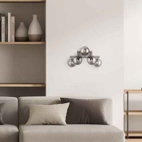 A minimalist living room features a beige sofa adorned with two cushions. Mounted on the wall is the Dallas 5 Light Wall Light in chrome with smoked mirrored glass. Wooden shelving units, decorated with vases and books, flank the sofa to enhance the decor.