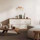 A minimalist living room includes a wooden coffee table, white couch, and modern dresser adorned with books, pottery, and a green vase. The Dallas 5 Light Wall Light in gold with amber mirrored glass casts a warm glow on the beige walls from above.