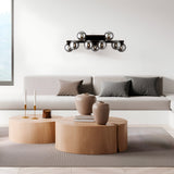 In a modern minimalist living room, a sleek sofa with gray cushions rests against a white wall. Two round wooden coffee tables feature decorative vases and candles. The Dallas 9 Light Wall Light - Black with Smoked Mirrored Glass enhances the contemporary lighting above.