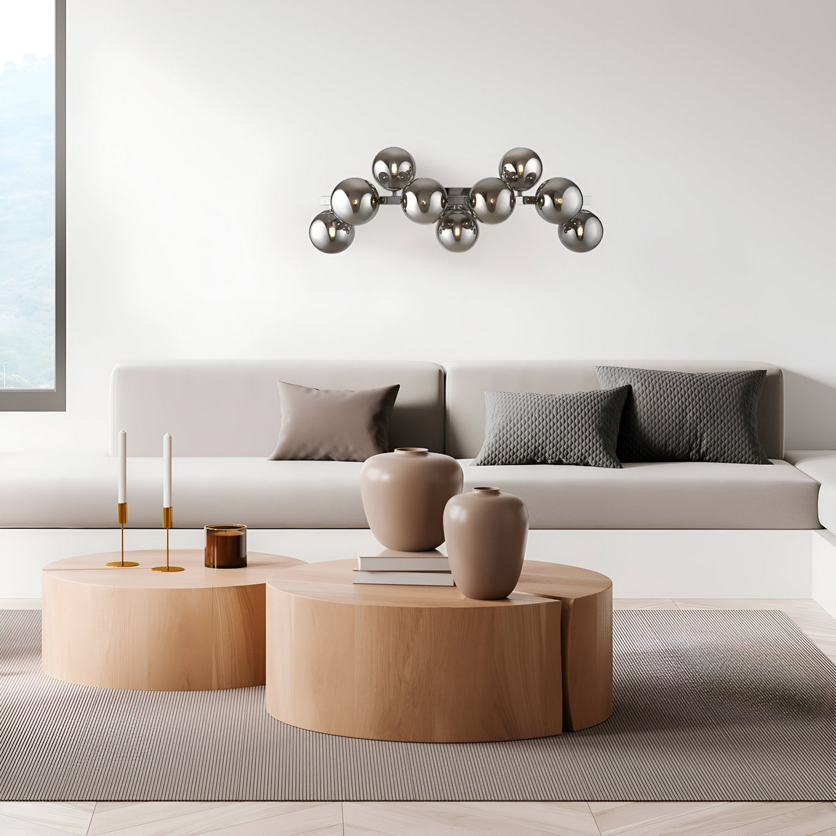 A modern living room features a light gray sofa with gray pillows. Two wooden coffee tables display neutral vases and candles. The Dallas 9 Light Wall Light with chrome and smoked mirrored glass adds elegance, complemented by a metallic wall sculpture and soft natural light from a large window.