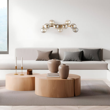 A modern living room features a minimalist beige sofa with gray pillows. Decorative vases and candles sit on wooden coffee tables, while a Dallas 9 Light Wall Light in gold with amber mirrored glass accentuates the white wall. To the left is a large window.