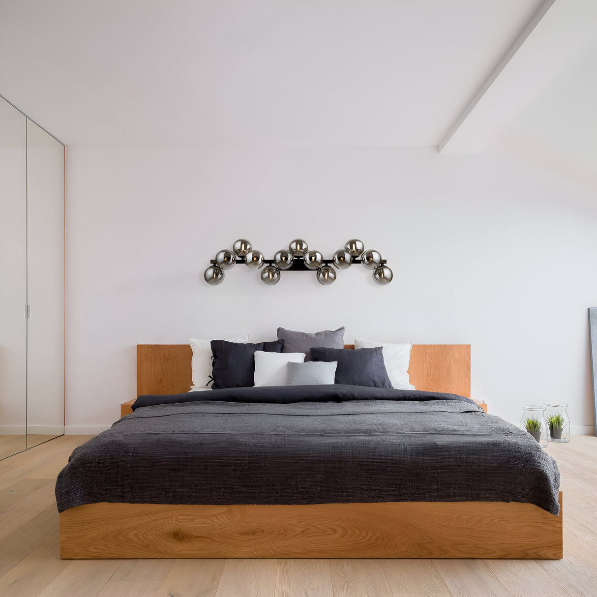 A minimalist bedroom features a wooden bed frame with gray bedding and pillows. A modern metal wall sculpture adorns the white wall above, complemented by the Dallas 13 Light Wall Light in black with smoked mirrored glass. The room boasts wooden flooring, adding an industrial touch.