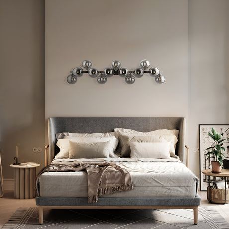 A modern bedroom showcases a gray upholstered bed with white and beige pillows and a brown throw. Above it, a geometric art piece pairs beautifully with the Dallas 13 Light Wall Light - Chrome With Smoked Mirrored Glass. A plant sits on the right, while a side table and candle are placed on the left.