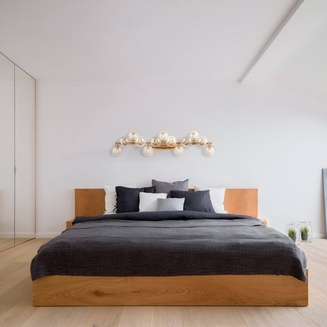 A minimalist bedroom showcases a wooden platform bed with gray bedding and pillows in white, gray, and black. Above the bed, a Dallas 13 Light Wall Light in gold with amber mirrored glass illuminates the space. The room features light wood flooring and a mirrored closet.
.