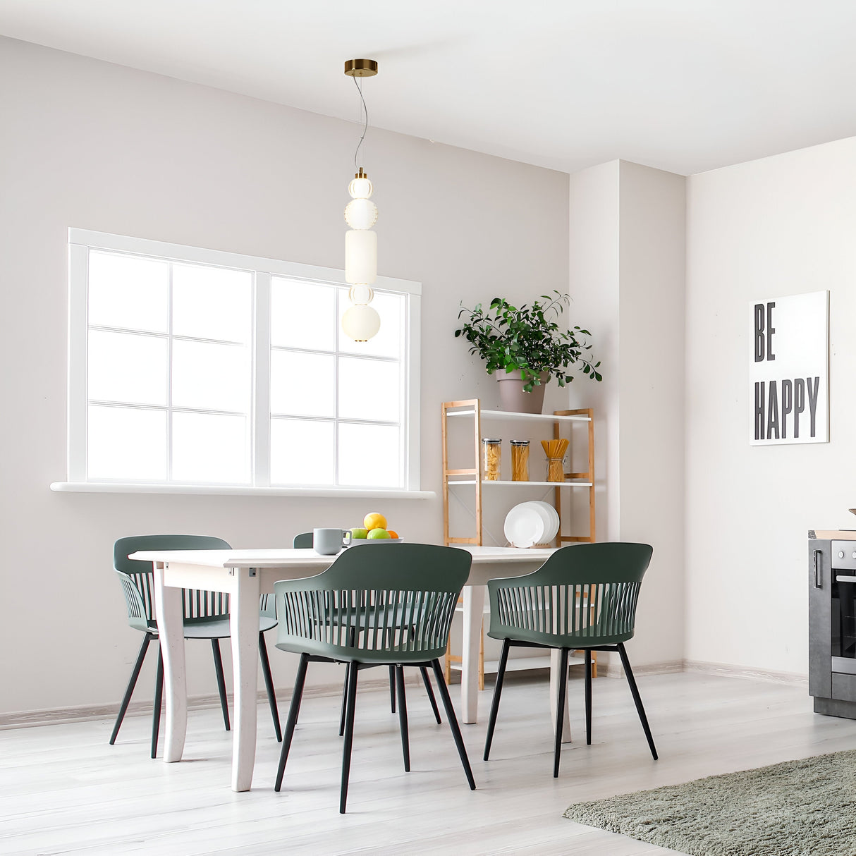 A bright dining area features a white table with four dark green chairs. A large window illuminates the wood shelf with plants and kitchen items. The Collar LED Pendant Light 35W - Gold complements the decor overhead, while a gray rug partially covers the light wooden floor.