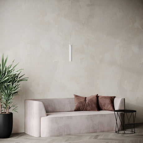 A minimalist living room showcases a light gray sofa adorned with two brown pillows, paired with a round black side table and a green potted plant. The Focus Single Pendant Light in white elegantly hangs from the ceiling against a textured, neutral-toned wall, exemplifying modern decor.