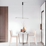 A minimalist dining area features a small white table with two chairs, a tall branch in a vase, two cups, and a copper jug. Above, the Supersymmetry LED Pendant Light 17W 4000K in Black enhances the modern aesthetics of this bright space perfectly.