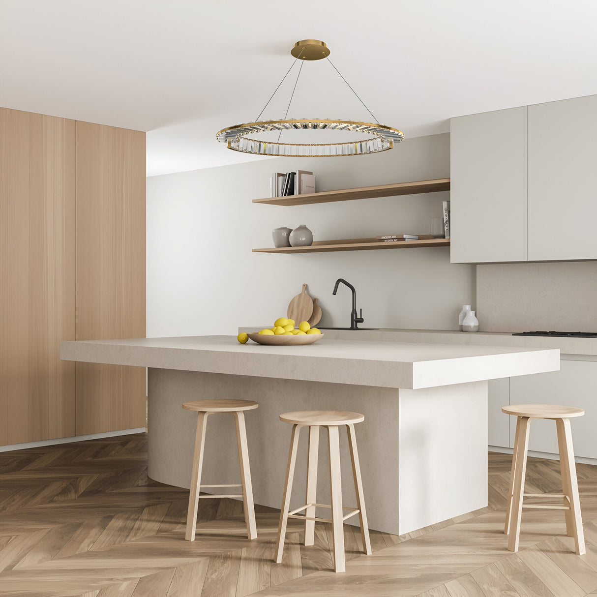 A modern kitchen includes a large white island with three wooden stools and a bowl of lemons. Light wood paneling contrasts with white cabinets and beige walls, while the Krone LED Circular Crystal Suspension Light 62cm - Gold elegantly hangs above. Open shelves display decorative items.