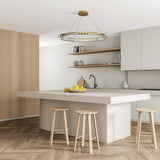 A modern kitchen includes a large white island with three wooden stools and a bowl of lemons. Light wood paneling contrasts with white cabinets and beige walls, while the Krone LED Circular Crystal Suspension Light 62cm - Gold elegantly hangs above. Open shelves display decorative items.