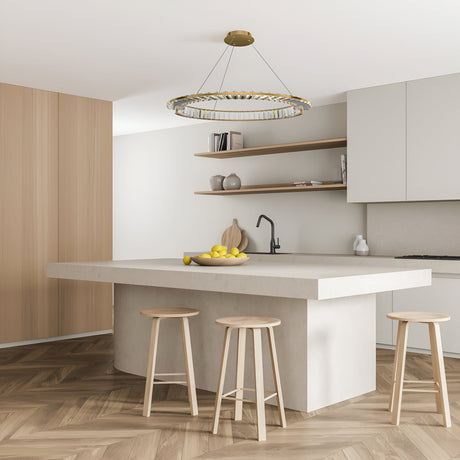 A modern kitchen includes a large white island with three wooden stools and a bowl of lemons. Light wood paneling contrasts with white cabinets and beige walls, while the Krone LED Circular Crystal Suspension Light 62cm - Gold elegantly hangs above. Open shelves display decorative items.