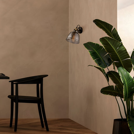 A minimalistic room with a wooden floor includes a black chair and an Irving Wall Light - Black With Smoky Glass. A green potted plant in the corner adds nature to the modern industrial decor.