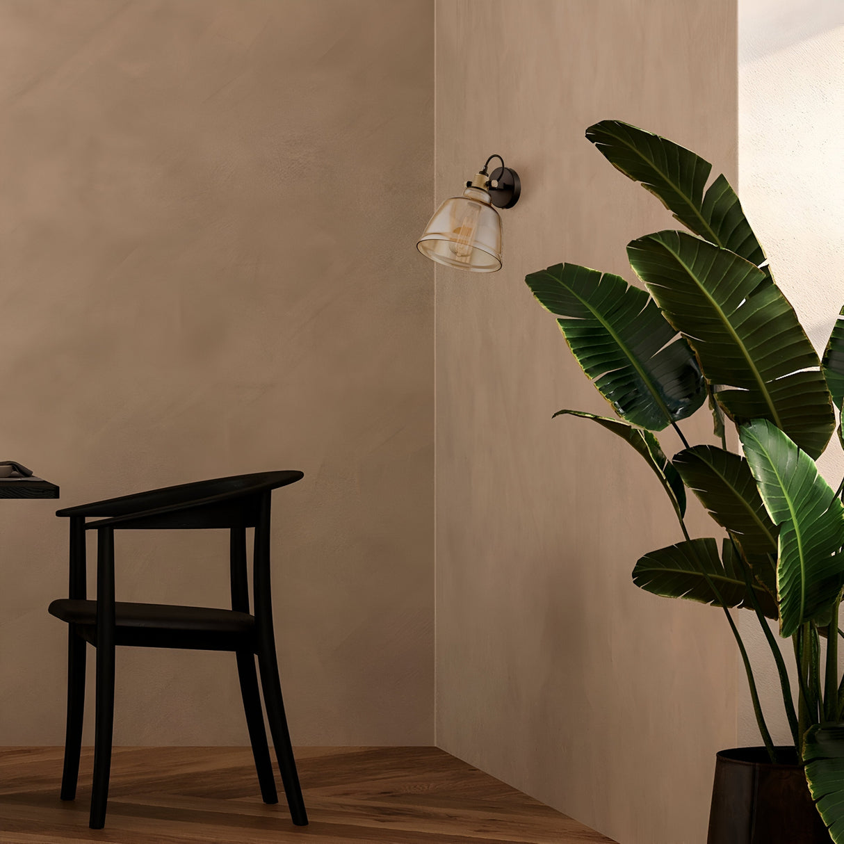 A minimalist room includes a black chair, a potted green plant in the corner, and the Irving Wall Light - Black With Amber Glass on the textured beige wall. Sunlight creates soft shadows on the floor, enhancing the serene ambiance.