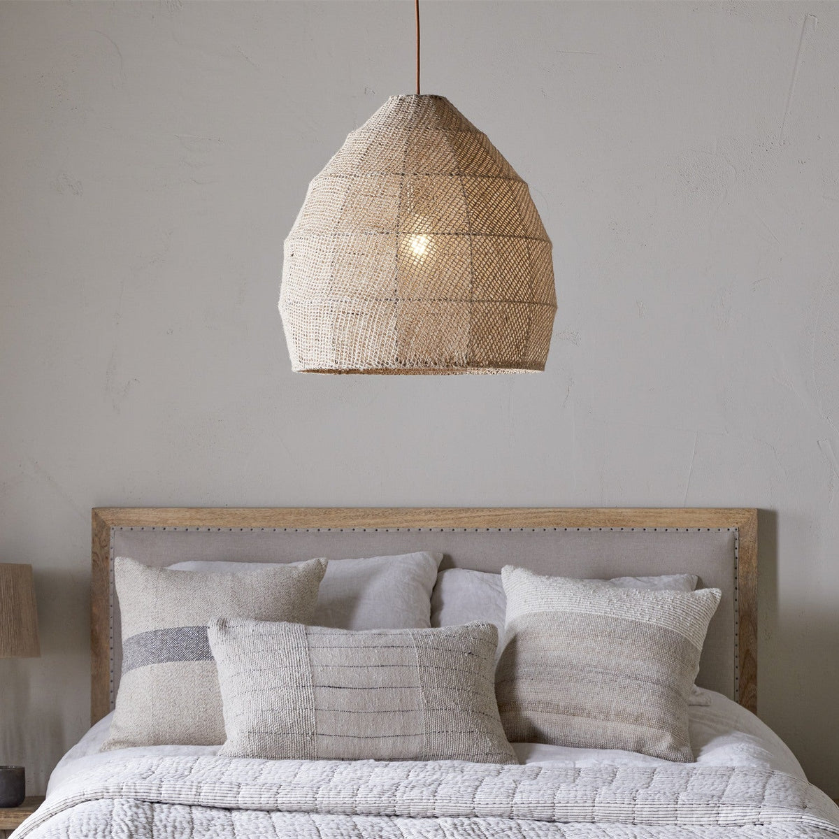 Jahari Woven Jute Pendant Light in natural jute with a domed, open-weave shade, shown hanging above a bed with linen cushions and a wood-framed headboard.