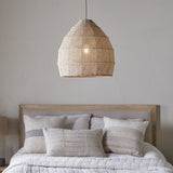 Jahari Woven Jute Pendant Light in natural jute with a domed, open-weave shade, shown hanging above a bed with linen cushions and a wood-framed headboard.