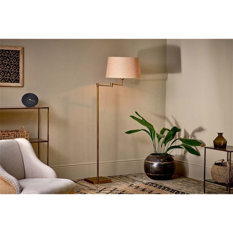 Kara Iron Floor Lamp Base in brass with swing arm and square base, shown with linen shade in a living room beside a chair and plant.