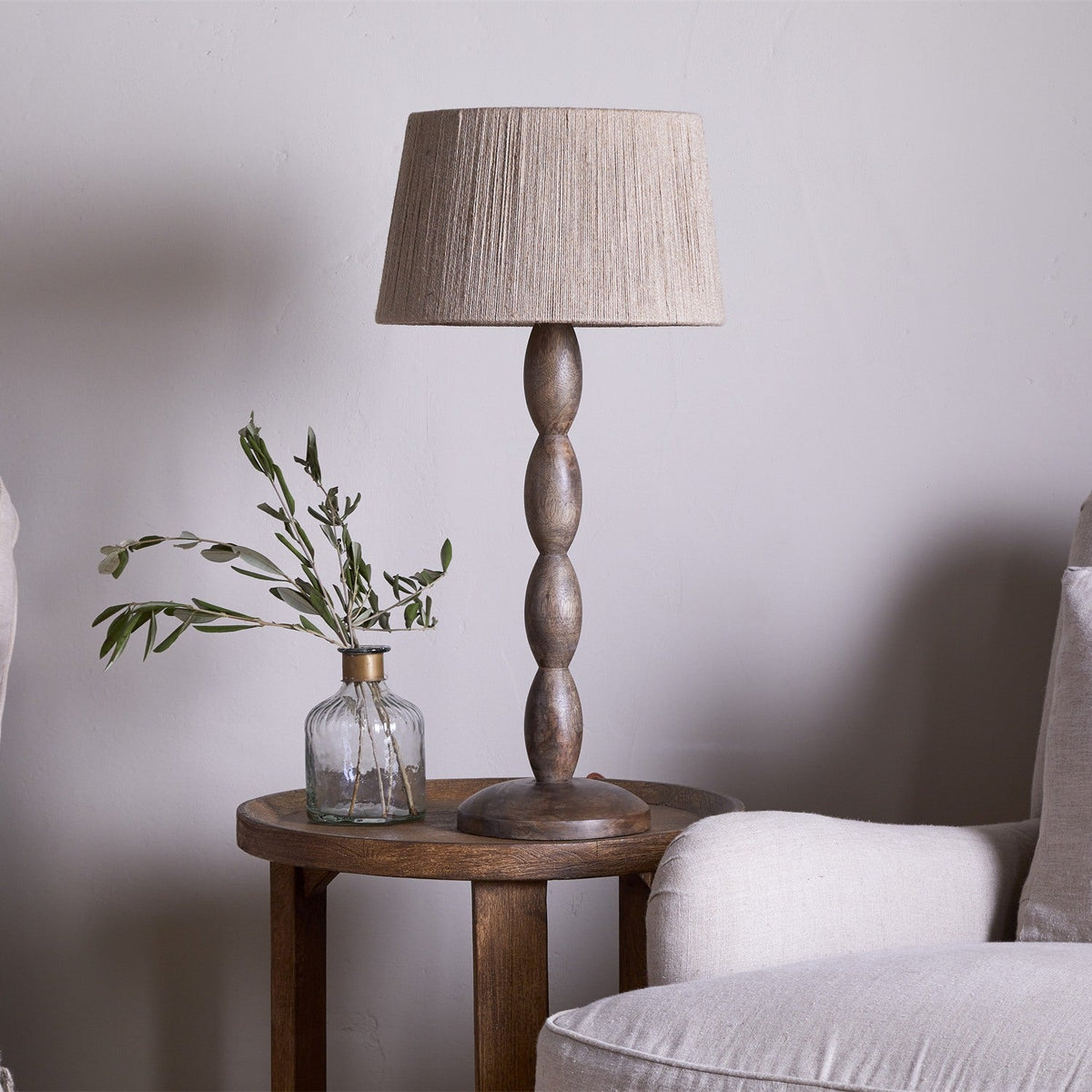 Konali Mango Wood Table Lamp with beaded, stacked-spindle column in ash stain and a tapered beige fabric shade, shown on a wooden side table beside a glass bud vase in a living room setting.