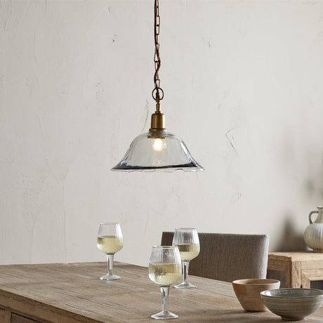 Keju Recycled Glass Pendant Light in Clear, a wide shallow bell shade with a rippled rim on a brass collar and chain, hung above a wooden dining table.
