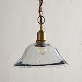 Keju Recycled Glass Pendant Light in Clear, a wide shallow bell-shaped shade with a rippled rim on a brass collar and stem, hung from a brass chain with rust fabric flex.