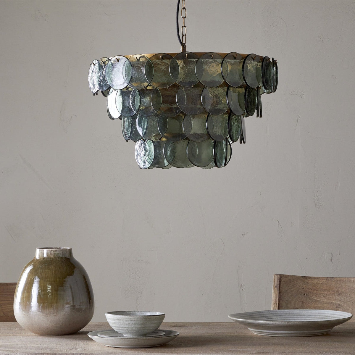 Kagitha Large 3 Light Recycled Glass Chandelier in Green, tiered round glass discs on a brass frame, hung in a dining room above a table with ceramic tableware.