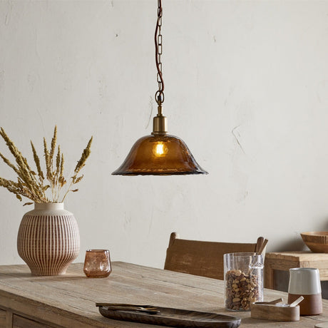 Keju Recycled Glass Pendant Light in Dark Amber with a wide, shallow rippled bell shade on a brass collar and chain, shown lit above a wooden dining table.