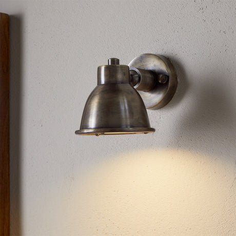 Kasai Brass Wall Spotlight in antique brass, mounted on a plain wall with its domed, adjustable head angled downward and switched on, casting a warm glow below.