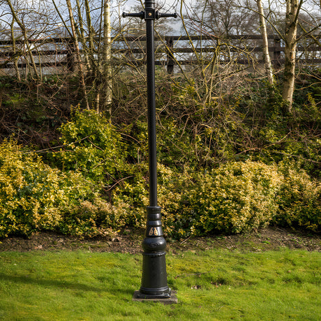 Victorian Cast Iron Lamp Post Column, 2.36m black metal pole with decorative base and cross-arm top, set on grass before hedges and trees in a garden setting.