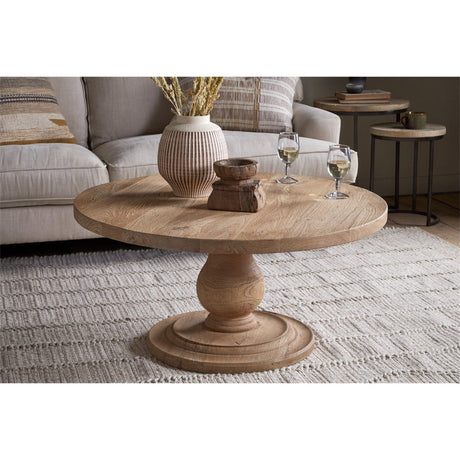 Laya Round Carved Mango Wood Coffee Table in natural wood with turned pedestal base, shown in a living room beside a sofa, side tables, and wine glasses.