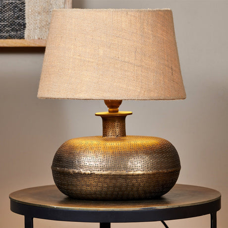 Lumbu Small Lamp Base in antique brass with a punched dot texture and rounded, flattened body, topped with a tapered linen lampshade on a round side table.