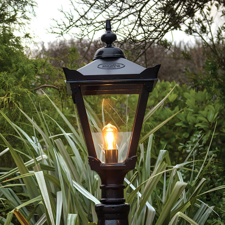 Victorian Manor black lantern head with 77mm fitting, glass panels, and lit bulb, mounted outdoors amid greenery.