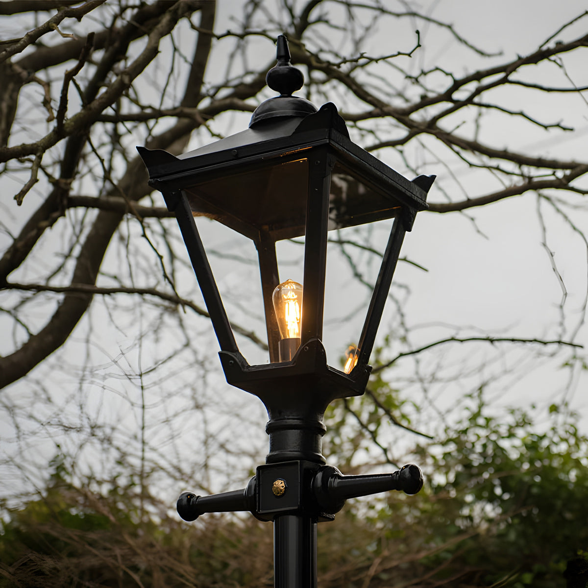 Victorian Manor black metal replacement lantern head with glass panels and lit bulb, mounted on a post, shown outdoors against bare branches.