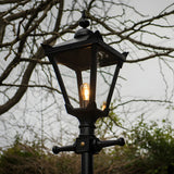 Victorian Manor black metal replacement lantern head with glass panels and lit bulb, mounted on a post, shown outdoors against bare branches.