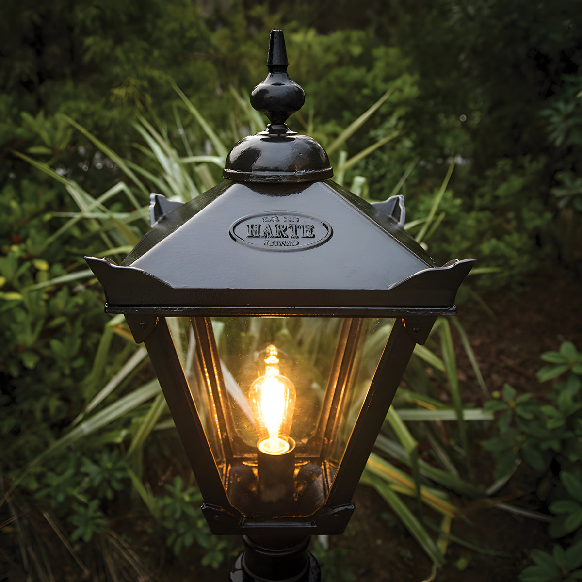 Victorian Manor black replacement lantern head with hexagonal glass panels and finial top, lit bulb inside, mounted outdoors against garden foliage.