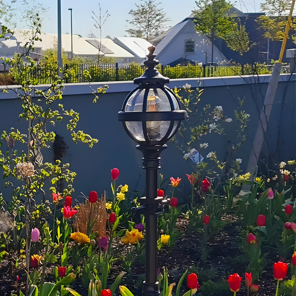 Black cast iron globe lantern head on a garden lamp post with clear glass panels, shown outdoors amid tulips and a garden wall.