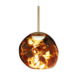 The Lava Small LED Pendant Light - Gold features an irregular, glossy glass shade with amber, gold, and dark red swirls, suspended from a slim brass rod and an adjustable cable for customizable hanging height.