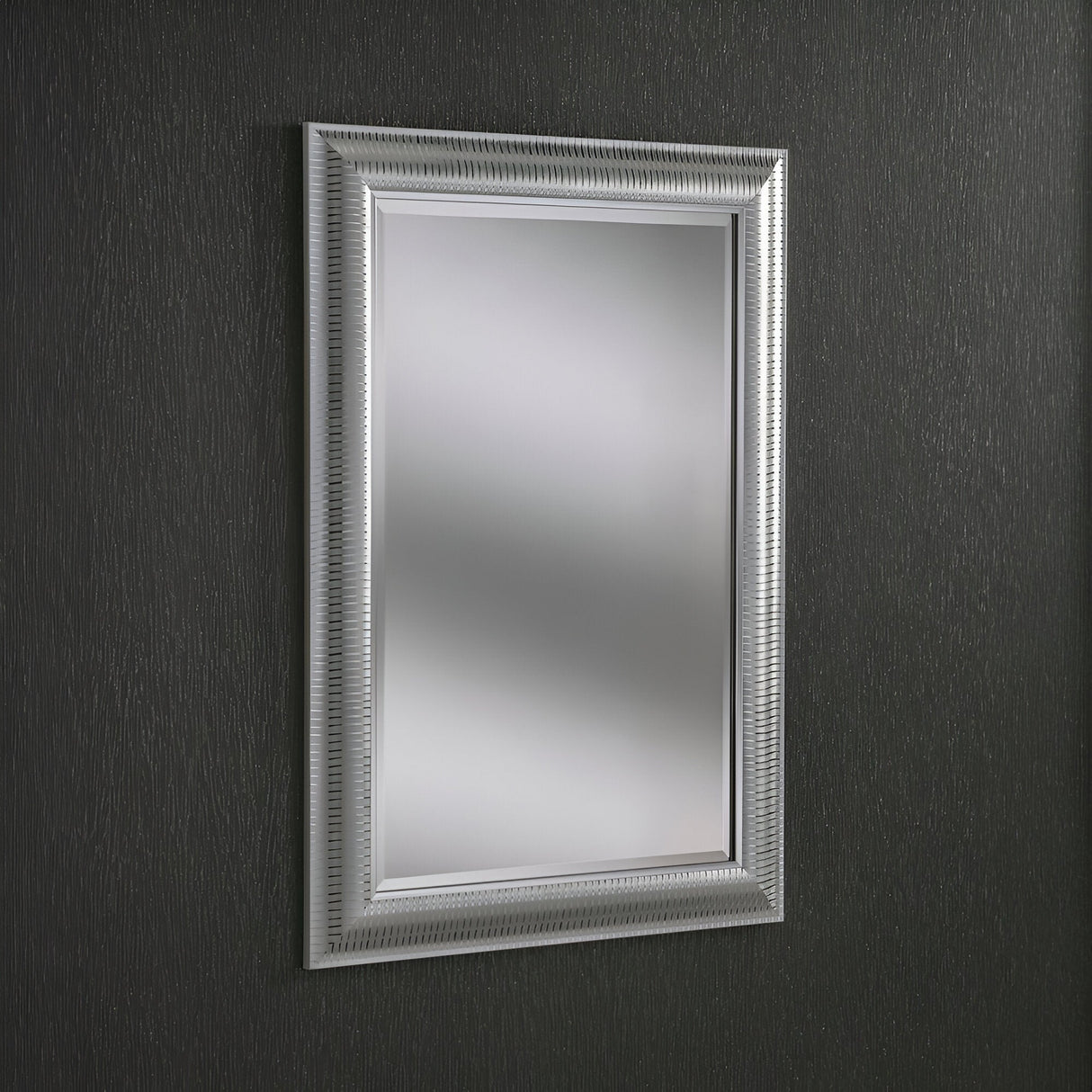 The Veris Rectangle Wall Mirror - Silver, featuring a textured silver frame, hangs on a dark, textured wall. Its intricate design adds elegance to the minimalist setting, while the dark tone of the wall mirror perfectly contrasts with its reflective surface.