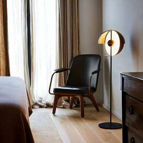 A modern bedroom showcases a black armchair beside the Lucia LED Floor Lamp - Black with a round handblown glass shade, casting warm light. Sheer curtains drape a tall window, and the edge of a bed and dresser are in view.