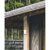 Kandanchu 2 Light round outdoor wall light in sand white aluminium, mounted on a timber post beside a dark doorway under a tiled eave.
