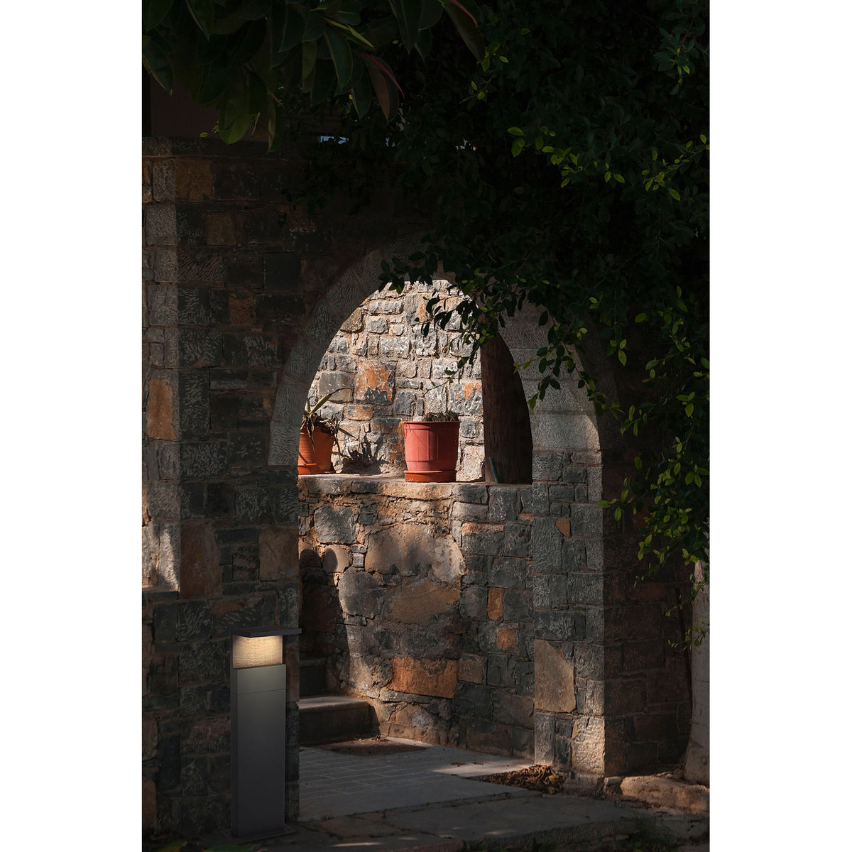 Ruka Floor Lamp in anthracite, a compact 60cm bollard-style outdoor light with a warm white glow, shown illuminating a stone archway pathway at dusk.