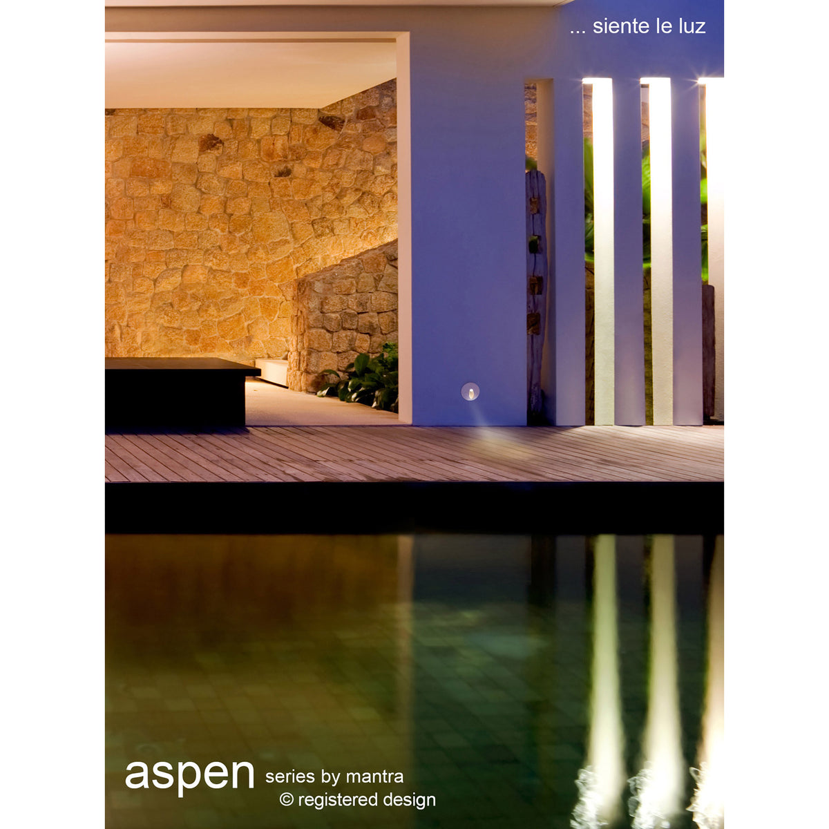 Aspen IP65 round recessed outdoor wall light in sand white, shown installed in a wall beside a decking area, casting a downward beam near a reflecting pool at night.