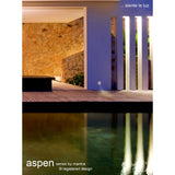 Aspen IP65 round recessed outdoor wall light in sand white, shown installed in a wall beside a decking area, casting a downward beam near a reflecting pool at night.