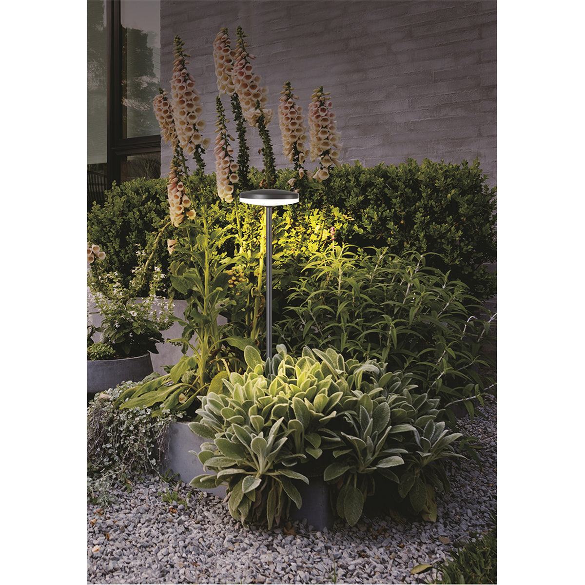 Akita IP54 Bollard Light, 60cm sand black aluminium post with round LED head, shown illuminating a planted garden bed beside a stone wall.