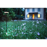 Akita IP54 Bollard Light, 80cm sand black outdoor post with round LED top, glowing warm white among wildflowers on a lawn near a lit house at dusk.