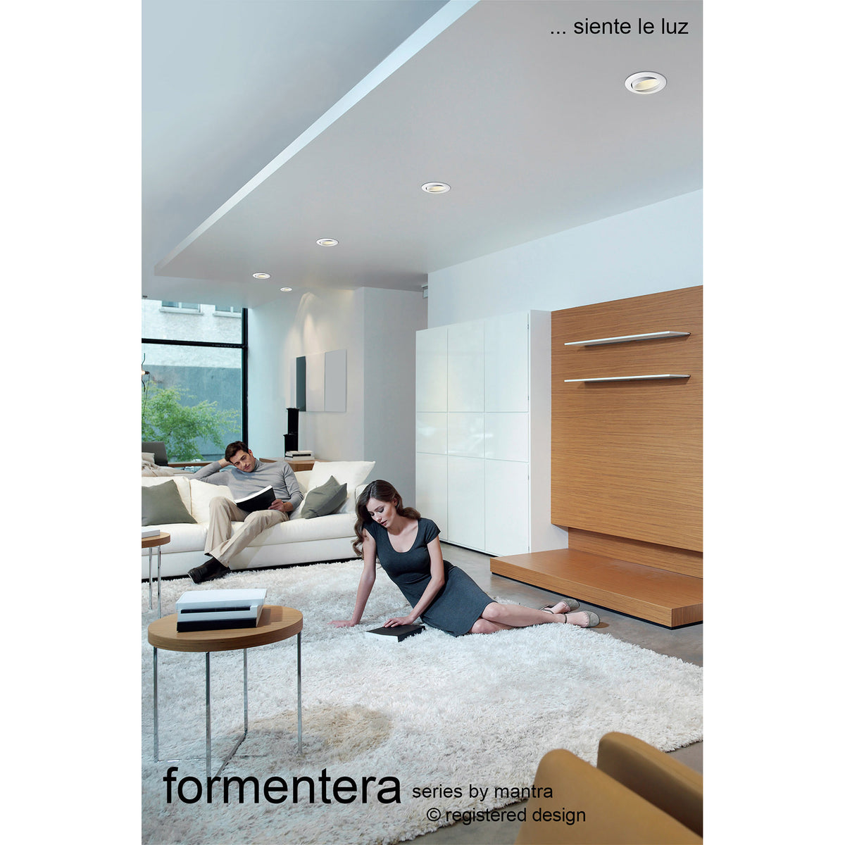 Formentera recessed downlights with matt white bezels installed in a living room ceiling, illuminating a neutral interior with sofa, rug and wood-panelled wall.