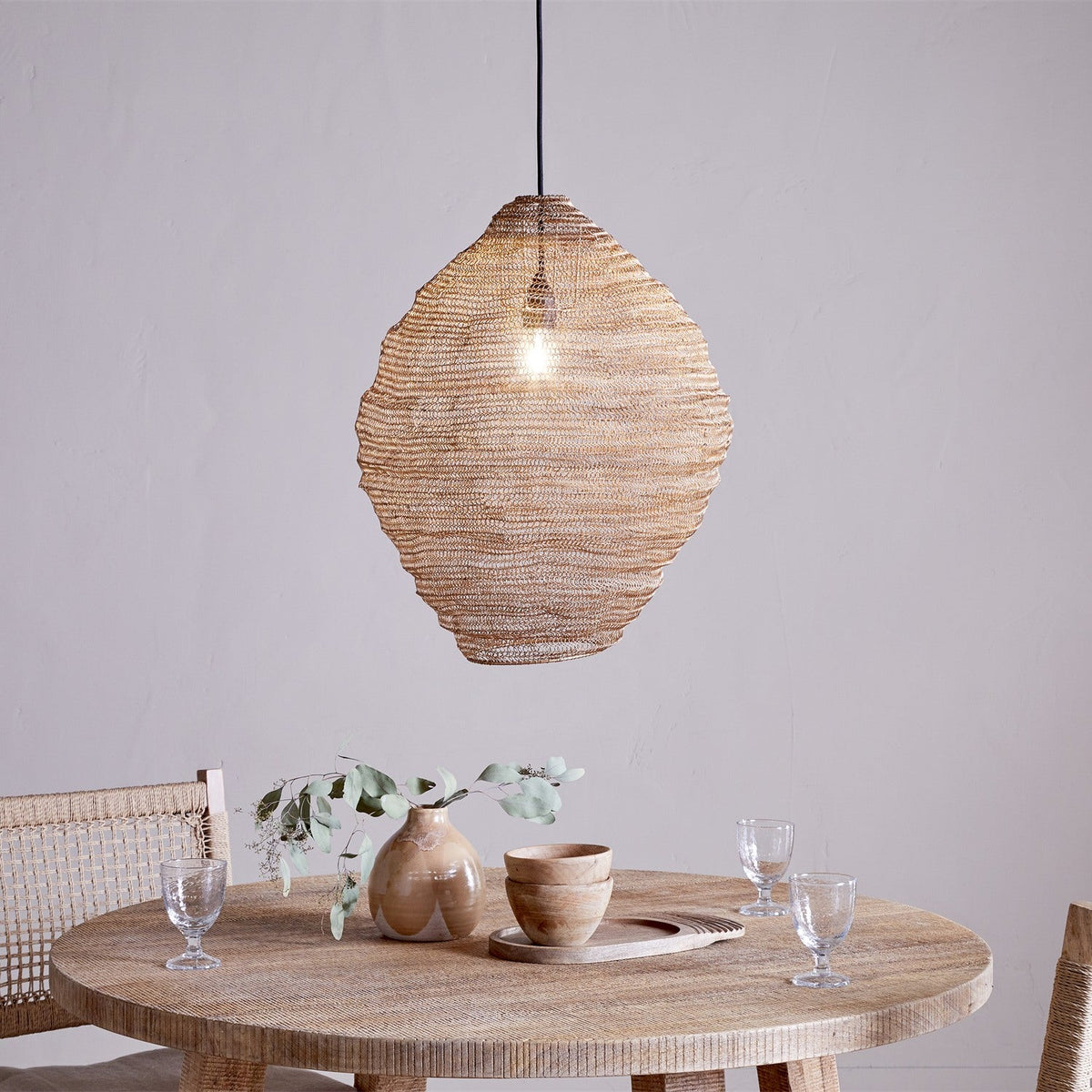 Mukuni Large Wire Lampshade in brass-toned knitted mesh, lantern-shaped with a full belly, hung as a pendant light above a round wooden dining table.