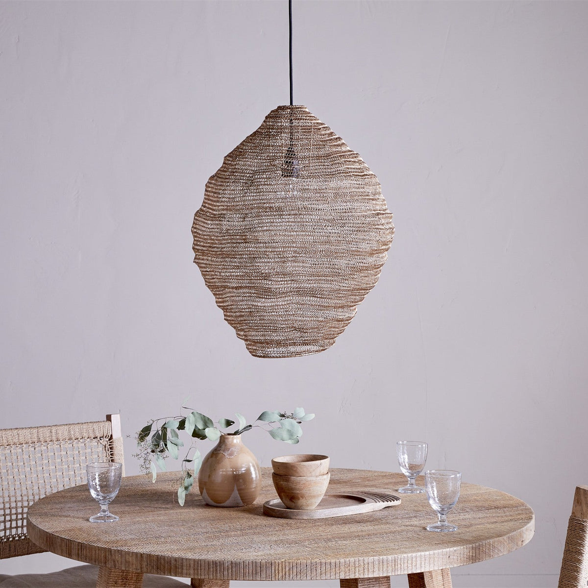 Mukuni Large Wire Lampshade in brass-toned knitted mesh, pendant style with a lantern silhouette, shown above a wooden dining table with tableware and a vase.