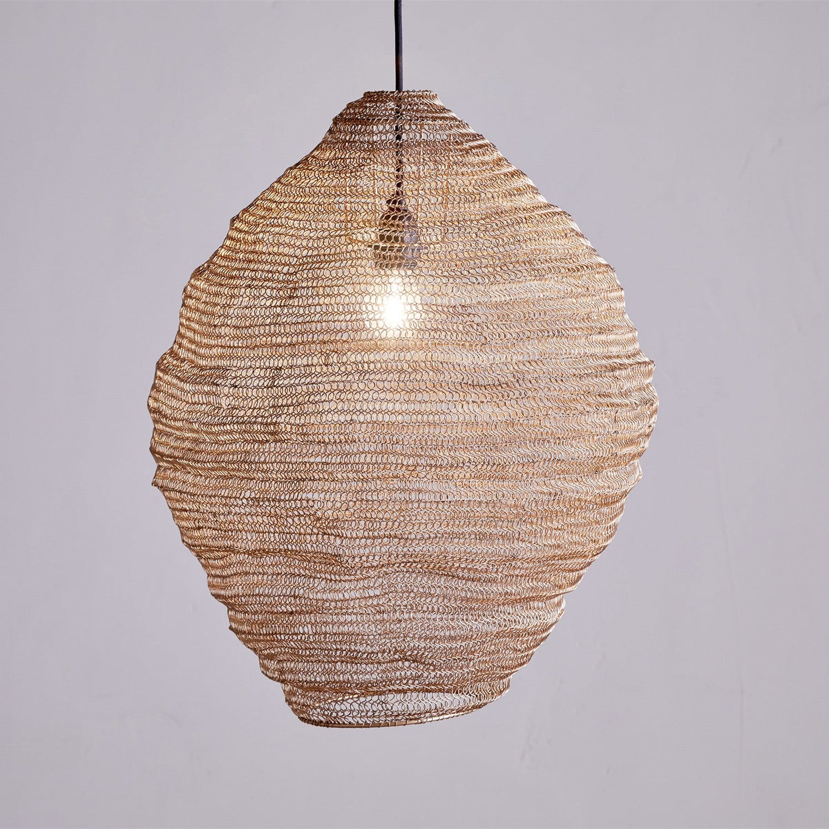 Mukuni Large Wire Lampshade in brass-toned knitted mesh, tall lantern shape with full belly, hung pendant-style with visible bulb against a plain grey background.