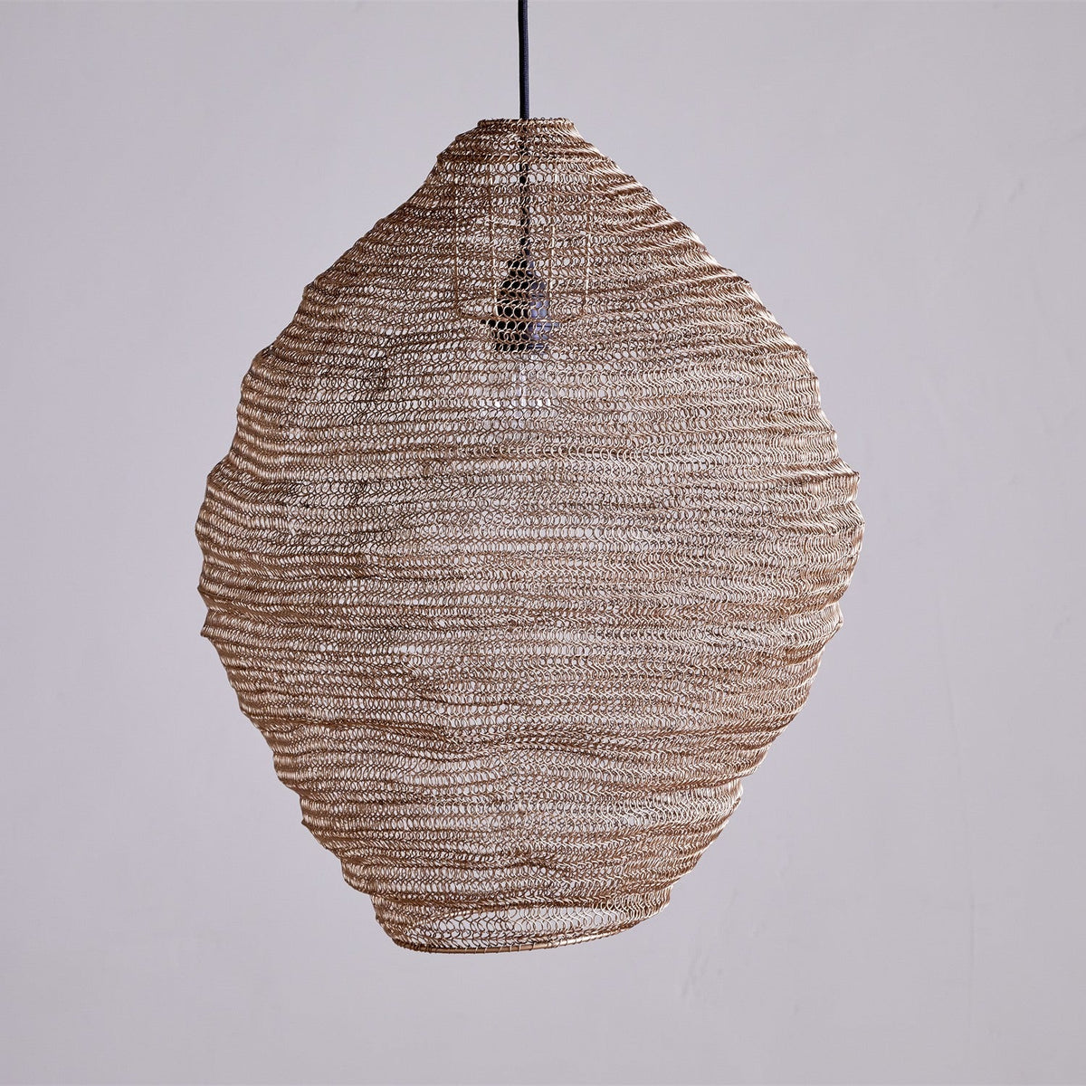 Mukuni Large Wire Lampshade in brass-toned knitted mesh, a tall lantern shape with a full belly tapering at the waist, hung against a plain light grey background.